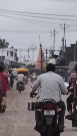 A view of a dirt road in India, with a temple visible in the distance. Several people are riding motorcycles on the road, and there are a few cars and carts as well. The sky is overcast.