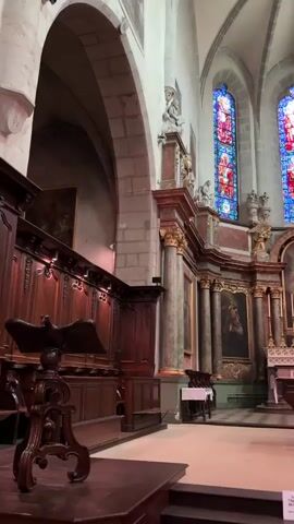 The video shows an interior view of a church. In the foreground on the left is a wooden lectern with an eagle-shaped stand. Behind it are rows of dark wooden pews with ornate carvings. The architecture features stone arches and columns. On the right, a man in a blue shirt and backpack is looking at a map or brochure. The background shows an altar area with a white altar, crosses, and ornate decorations. Above the altar are stained-glass windows with vibrant red, blue, and white figures. The ceiling is vaulted and appears to be made of stone. Paintings adorn the walls. The lighting is dim, with light coming from the stained-glass windows and some artificial sources. The overall atmosphere is solemn and majestic.