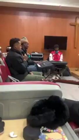 The video shows three young black men rehearsing a musical performance in a room that looks like a church or chapel. The room has wood-paneled walls, a cross on the wall, and a television screen. Two of the men are playing keyboards, one wearing a black beanie, and the other a red jacket and black hat. The third man, in a black jacket, is singing into a microphone. The video is shot from the perspective of someone holding a guitar, and you can see a fluffy black item, rosary beads, and an earbud case on a table in the foreground.