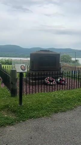 The video shows Uzal Knapp’s gravesite. The tombstone is dark gray, rectangular, and sits on a raised, square base. It is surrounded by a black wrought-iron fence, with decorative wreaths at its base. A small informational sign stands next to it, with a picture of Uzal Knapp. Behind the site, there is a chain-link fence and a green lawn. Beyond that, in the background, there is water and mountains. The weather appears cloudy.