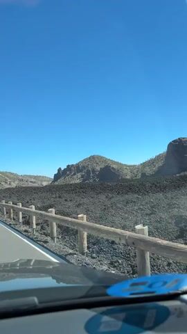 The video shows a view from a vehicle driving along a road. The road is paved and has white lane markings. A wooden fence with metal posts runs alongside the road. Beyond the fence is a vast landscape of dark, rocky terrain, possibly volcanic rock. In the distance, there are mountains with rugged peaks and sparse vegetation. The sky is clear and bright blue. The camera perspective suggests the video is shot from the dashboard or windshield area of the vehicle, indicating a forward-facing view of the road and the surrounding environment. There are no people visible in the video, and the only observable objects are the road, fence, mountains, and sky. The overall scene depicts a scenic drive through a remote, natural landscape under a clear, sunny sky.
