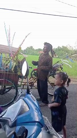 The video depicts a man in traditional Javanese attire standing beside a decorated bicycle rickshaw, accompanied by a young child with pigtails. The man wears a striped shirt, black pants, and a traditional headdress. The child, standing with arms akimbo, is dressed in a dark-colored outfit with a floral pattern. The rickshaw behind them is adorned with colorful decorations.

In the foreground, partially obscuring the view, is a blue scooter. The setting appears to be a rural area, with a green rice field visible in the background under a cloudy sky. The scene is captured from a static camera position.