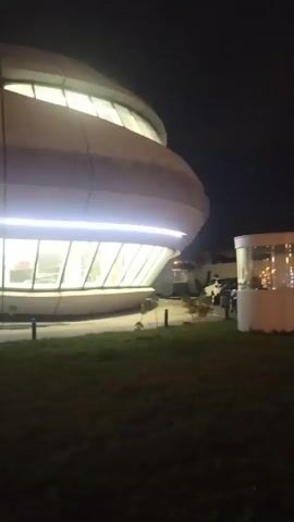 At night, the camera slowly pans from right to left across the exterior of a modern building with a curved, multi-tiered facade lit by white lights. An SUV is parked on the grass next to the building's entrance. The background is dark.