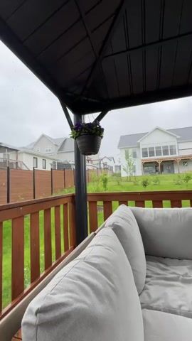 The video shows a view from a covered patio looking out at a backyard on a cloudy day. In the foreground, a large, light-colored sectional couch is visible. A dark-colored gazebo roof extends over the patio. A hanging flower basket with purple flowers is suspended from the gazebo. A wooden railing borders the patio. Beyond the railing, a lawn stretches out with some young trees. In the middle ground, there are several modern houses with white siding and dark roofs. A wooden fence runs along the property line. The sky is overcast, suggesting rainy or gloomy weather. The video captures light rain or water droplets falling.