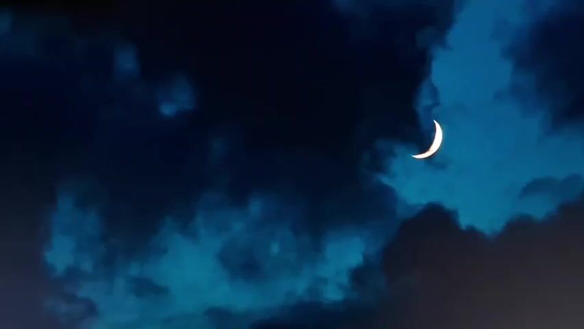 A crescent moon is visible in the dark blue sky, partially obscured by dark, swirling clouds. The moon is bright and appears to be reflecting light. The clouds are varied in texture and density, with some areas appearing more opaque than others. The overall impression is of a cloudy night sky with a clear, thin moon.