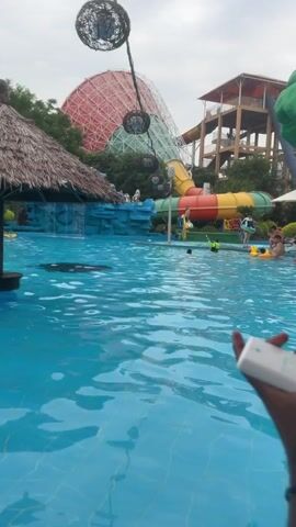 A video shows a pool at a water park. In the foreground, a person's hands are visible, holding a white object. In the middle ground, the blue water of the pool fills the frame, with several people swimming and playing. Some are wearing life vests. In the background, a large, colorful water slide complex is visible. One slide is yellow and red, another is green, and there is a large geodesic dome structure behind the slides. The sky is overcast.