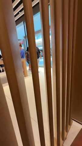 The video shows the interior of a store, with a wooden slatted divider in the foreground. Through the slats, several people are visible. On the left, a person in a blue shirt is standing behind a counter or display. To their right, a person wearing a black shirt and white shorts is looking down at something. Further in the background, a person in a striped shirt and denim shorts walks away from the camera. Another person, wearing a dark shirt and light-colored pants, walks towards a glass door in the background. The environment appears to be a brightly lit retail space.