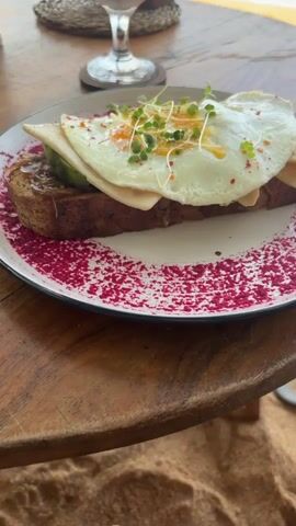 The video begins with a close-up shot of a breakfast meal consisting of a slice of brown toast, topped with a spread, avocado, cheese, and a fried egg, garnished with microgreens on a white plate decorated with a red pattern.  A beverage in a stemmed glass sits behind it on a wooden table. The camera zooms out to reveal a beach setting with boats in the distance, the ocean, and the sky with clouds. A sign displays menu items. The camera continues to move, revealing lounge seating on the beach.