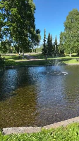 The media shows a tranquil park scene with a pond and a fountain. The water in the pond is clear, revealing the pebbly bottom. A fountain sprays water in the center of the pond, creating ripples on the surface. Lush green trees surround the pond, their leaves rustling gently in the breeze. The sky is a brilliant blue, dotted with a few wispy clouds, suggesting a pleasant day. In the background, a path winds through the park, and a couple of benches are visible, hinting at a place for relaxation and contemplation. The overall ambiance is one of peace and natural beauty. The time of day appears to be late morning or early afternoon, as indicated by the bright sunlight casting distinct shadows. While no people are prominently featured, the presence of benches suggests human activity in the area. The scene evokes a sense of calm and serenity.