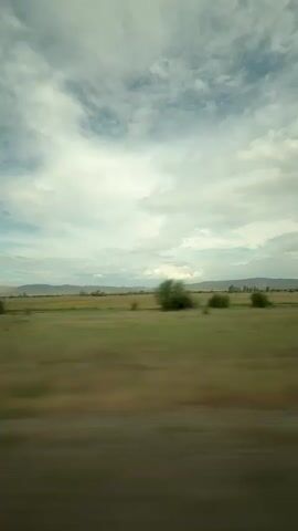 This is a landscape video taken from a moving vehicle. The video shows a vast, open field with scattered trees and a distant mountain range under a cloudy sky. The motion blur indicates that the camera is moving at a significant speed. The sky is a mix of white clouds and patches of blue sky, suggesting it might be daytime. The field appears to be covered in green and brown vegetation, likely grass or crops. The trees are small to medium-sized and appear to be deciduous. The overall impression is of a rural or natural landscape. There are no people or man-made structures clearly visible in the frame.
