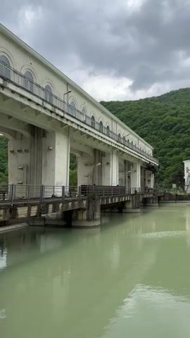 The video shows a large, white concrete structure over a body of water. The structure has multiple archways along its upper section and appears to be a dam or part of a hydroelectric plant. The water is a pale green color.  Behind the structure is a forested mountain under a cloudy sky. To the right, there is another smaller white building with similar architectural features. The video seems to be taken from a stationary position, and there is no evident movement.
