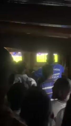 A group of people are gathered in a room watching a soccer game on two televisions. Some are wearing jerseys with the word "INTERSC" visible on one. Some are taking photos or videos of the TVs or of the crowd. The lighting is dim. The ceiling appears to be made of wood.  The crowd is cheering and celebrating.