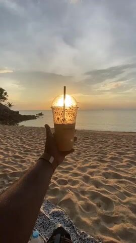 The video shows a person's arm and hand holding a plastic cup with a straw, against a beach scene at sunset. The sun is positioned behind the cup, creating a backlit effect. The beach has tan sand with scattered footprints. The sea is calm with a hint of waves, and the horizon is slightly obscured by clouds. A rocky coastline is visible to the left with some vegetation. In the distance, a person is walking along the shore. At the bottom of the frame, there's a glimpse of patterned fabric and a water bottle, suggesting the person is sitting or resting on the beach.