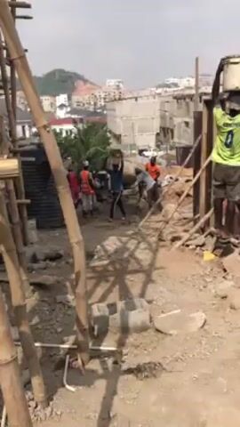 The video shows a construction site on a sunny day. Several construction workers are visible, some carrying buckets and others carrying materials on their heads. There's a concrete mixer in the background. The site has scaffolding made of bamboo and wooden forms for construction. In the distance, there are buildings and a hillside. The ground is dirt and gravel.  Workers are walking around the site and moving materials.