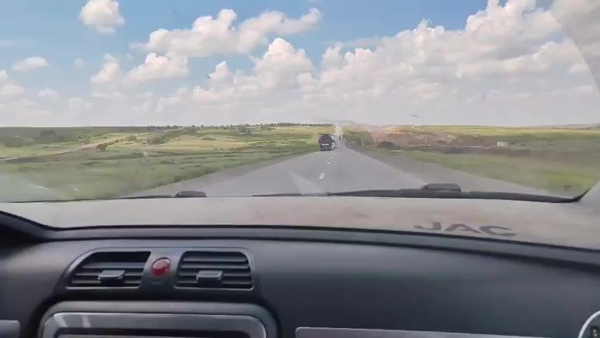 This video provides a first-person perspective from inside a vehicle, looking through the windshield. The vehicle is traveling on a paved road through a vast, open landscape. The road is bordered by grassy fields and some distant hills. The sky is blue with scattered white clouds. Several vehicles are visible on the road ahead. Initially, there are several trucks in the distance. As the vehicle progresses, a white truck passes by on the left. Further down the road, there are smaller cars. The interior of the vehicle is visible in the foreground, showing part of the dashboard, air vents, and a rearview mirror. The dashboard has the "JAC" logo. The overall impression is a journey through a rural or semi-rural area on a clear day.