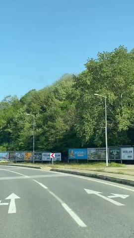 The video shows a first-person perspective from a vehicle driving on a paved road.  The sky is clear and blue.  To the right of the road, a tree-covered hill rises. Along the roadside, a series of billboards are visible, displaying advertisements.  As the vehicle advances, road signs come into view, indicating directions to AĞVA, ŞEHİR MERKEZİ, Ahmetli, Kandıra, KOCAELI, and ŞAKLIGÖL. Other vehicles are visible on the road ahead.  The road has painted white lines marking lanes and intersections. Streetlights line the road.