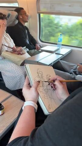 The video shows a first-person perspective of someone drawing in a sketchbook on a train. The drawer's hands are in the foreground, holding a pen and the sketchbook, which contains a line drawing of two faces. The setting is inside a train carriage with windows on the right side showing a blurred outdoor scene. In the background, there are other passengers, including a man wearing glasses and earbuds, and a woman sitting next to him. Several tables are visible, with laptops and a water bottle on one of them. The focus is on the drawing activity and the immediate surroundings within the train.