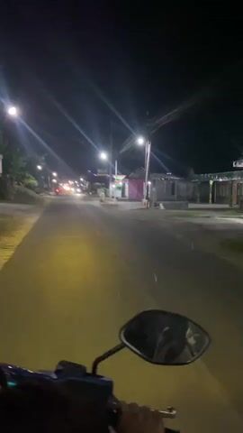 The video captures a first-person perspective of someone riding a motorcycle at night on a paved road. Streetlights illuminate the scene, casting yellow and white light. The road appears to be in a somewhat rural or suburban area, with low-lying buildings visible on either side. There are other vehicles on the road, including motorcycles and cars, identifiable by their headlights. The rider's hand is visible gripping the handlebars, and a rearview mirror is in the foreground, reflecting the dark sky and lights. The overall atmosphere is nocturnal and somewhat deserted, with the sounds of passing vehicles and the hum of the motorcycle implied.