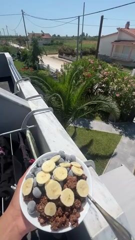 A close-up shot focuses on a white bowl filled with cereal, sliced bananas, and dark, round berries, held by a hand with a spoon. The background reveals a sun-drenched outdoor scene with a lush green lawn, blooming pink flowers, and a tall palm tree. In the distance, white and pink houses with red-tiled roofs are visible, along with a small boat. The sky is a clear, bright blue, indicating a warm, sunny day, likely in the morning or early afternoon given the lighting. The setting appears to be a residential area in Chaidefto, Greece, characterized by Mediterranean-style architecture and abundant vegetation. The overall impression is one of a peaceful, idyllic morning meal enjoyed outdoors.