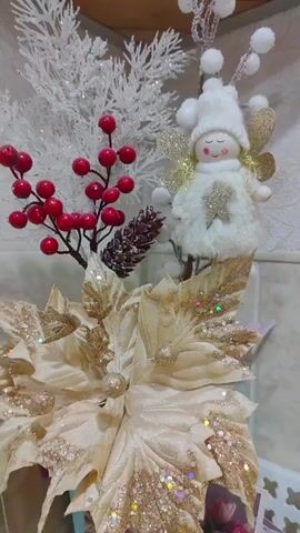 The video shows a close-up of a decorative arrangement. In the foreground, a large, cream-colored artificial poinsettia flower is visible. Its petals are adorned with gold glitter and scattered colorful sparkles. Behind the poinsettia, to the left, is a sprig of artificial red berries and a small pine cone. To the right, a small, fluffy white angel figurine wearing a hat and with gold glitter wings is positioned above the flower. The angel has a smiling face with closed eyes. Behind these elements, a white, frosted artificial branch with small white pom-poms and another white, frosted, tree-like decoration can be seen. The background appears to be a wall with a patterned surface, possibly tile. The lighting suggests an indoor setting.