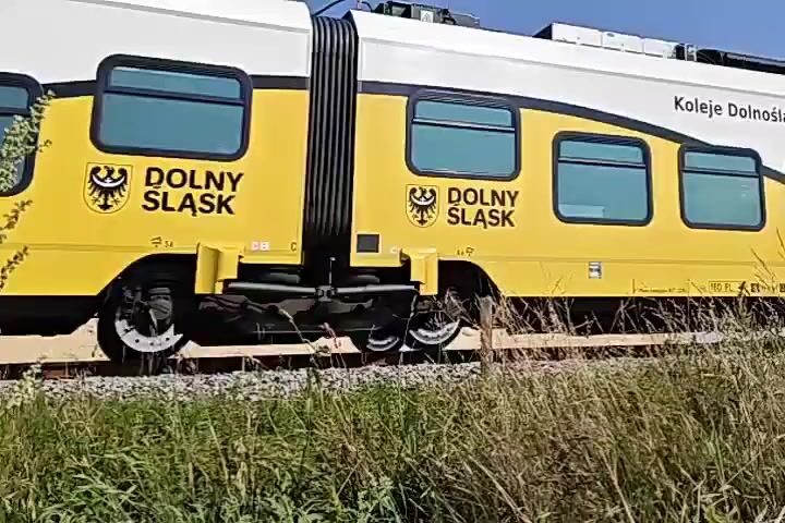The video begins with a close-up of a train car. The train is white with a yellow stripe along the bottom and features the letters "DKA" on its side. The train then moves away from the camera, revealing more of its exterior, which includes windows and some graphics. The train is on tracks surrounded by dry grass and vegetation. The camera pans to the right, showing a large pile of sand or dirt in the background. The sky is clear and blue, and there is a power line tower visible in the distance. The train continues to move forward and out of frame. The final shot shows the train in the distance, with the power line tower and construction materials in the foreground. The scene appears to be an outdoor environment, possibly a construction site or an area near railway tracks.