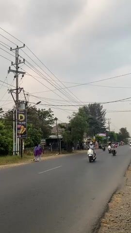 The video shows a street scene with moderate traffic. In the foreground, a white scooter with a person wearing a white helmet is moving from left to right. To the left of the road, there is a utility pole with multiple wires stretching across the frame, and a billboard advertising a product for 15,000. Two people in purple outfits are walking on the sidewalk near the billboard. Further down the road, several other scooters and motorcycles are visible, some moving away from the camera. The road is paved and appears to be a two-lane street with a shoulder on the right side. Trees and buildings line the side of the road, and the sky is overcast. The overall atmosphere is that of a typical day in a populated area with vehicular movement.
