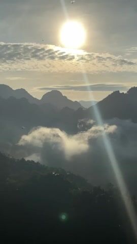 The video captures a serene mountain landscape at sunrise. The sun, a bright orb, is positioned high in the sky, casting long rays of light that stream across the frame. Wispy clouds dot the sky, adding texture to the dawn. Below, the rugged peaks of mountains recede into the distance, shrouded in a soft, ethereal mist that drifts through the valleys. The foreground is dominated by a dark, verdant slope, likely covered in dense forest, with hints of human structures or clearings barely visible through the shadows. The overall impression is one of tranquility and natural beauty, with the rising sun illuminating the majestic scenery.