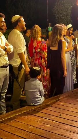 A group of people are gathered outdoors at night. The people are dressed in formal attire, including kippahs on some of the men, indicating a Jewish gathering. In the foreground, a young boy sits on the wooden deck. He is facing away from the camera. Behind him, a line of adults stand, some looking to their left, where a woman in a vibrant, floral dress is speaking. To the far left, two men in light-colored shirts and dress pants stand with their arms crossed. The environment appears to be a backyard or outdoor event space, with lush greenery visible in the background. The lighting suggests a celebratory or religious occasion.