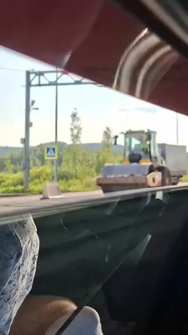 The video shows a view from inside a vehicle, looking out through the windshield and side window. In the distance, a large yellow road roller is compacting asphalt on a road. To the left of the road roller, a white truck with a grey trailer is parked. There are trees and vegetation in the background, and the sky is blue with some clouds. The camera is moving, as indicated by the slight blur in the foreground and the shifting perspective. The video captures a typical scene of road construction or maintenance.