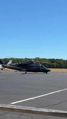 A dark-colored helicopter is parked on an asphalt tarmac under a clear blue sky. The helicopter has two main rotors and a smaller tail rotor. It has a sleek, modern design with windows along the side. There are red and white stripes on the fuselage near the tail. Another helicopter, also dark-colored, is visible in the background to the left. Trees and foliage form a treeline in the distance. The tarmac has white painted lines, and a portion of a wooden barrier is visible in the foreground. The scene appears to be an airport or helipad on a sunny day.