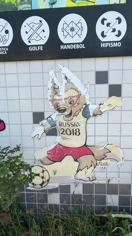 The video pans across a tiled wall decorated with cartoon characters and circular pictograms representing Olympic sports. The first character visible is a wolf-like mascot wearing a shirt that says "RUSSIA 2018", kicking a soccer ball. To its left, a pink and white cat-like mascot, identified as Miraitowa, the mascot for the Tokyo 2020 Olympics, is displayed. Further to the left, a red and pink character is partially visible. The pictograms above the wall include representations of gymnastics, golf, handball, hippism, fencing, trampoline, soccer, artistic gymnastics, boxing, climbing, cycling disciplines (BMX freestyle, BMX racing, road cycling, track cycling), and potentially other sports. Green foliage and grass are visible in the foreground, partially obscuring the lower portion of the wall and the mascots. The lighting suggests it is daytime, outdoors.