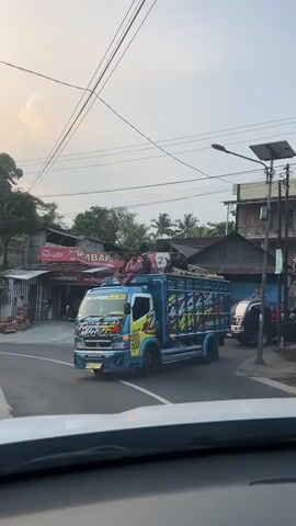 A blue and yellow truck is driving down a road. Several people are sitting on top of the truck's cargo area. The truck is decorated with various stickers and graphics. In the background, there are buildings and trees. The sky is overcast.