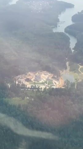 An aerial drone shot captures a sprawling construction site nestled within a lush, forested landscape. The site, dominated by light-colored structures, appears to be a complex of buildings, possibly a church or religious monument, indicated by the golden dome visible in the center. Surrounding the central construction area, the ground is a mix of disturbed earth, scaffolding, and partially completed structures, suggesting ongoing building activities. A small, dark lake sits to the right of the main site, with a winding path leading towards it from the construction zone. In the distance, a body of water, possibly a river or lake, is visible, bordered by a line of trees and a developed area with houses and what looks like a marina. The scene is hazy, with sunlight filtering through the atmosphere, creating soft glows and casting distinct rays of light across the landscape, particularly on the left side of the frame. The overall impression is one of a large-scale development project in a natural setting.