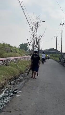 A person wearing a white hard hat and a black t-shirt walks along a paved road. They are carrying a shovel and a pickaxe. Several other people are walking or riding motorcycles in the distance on the same road. The road is lined with overgrown vegetation on the left and buildings in the distance on the right. The sky is overcast.