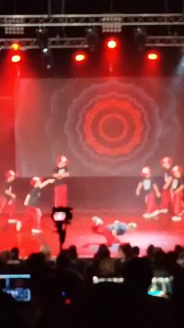 The video shows a dance performance taking place on a stage in a dimly lit venue, likely an auditorium or theater. The stage is set with a large screen displaying a geometric, mandala-like pattern in red and white. Several spotlights illuminate the stage, casting beams of light through the smoke or haze that fills the air.

The subjects are a group of young dancers, appearing to be children or pre-teens, dressed in matching outfits of black t-shirts and red pants, along with red baseball caps. They are engaged in a choreographed dance routine. One dancer is performing a move on the floor in the center of the stage, while the others are positioned around them, some in dynamic poses. The audience is visible in the foreground, silhouetted against the stage lights, with many people holding up phones to record the performance.

The event appears to be a dance recital or competition. The atmosphere seems energetic and focused, with the dancers concentrating on their performance and the audience actively observing and documenting the event. There is no visible text in the video. The lighting suggests an evening or indoor event, with no indication of weather conditions. The specific location within Mykolaiv, Ukraine is not discernible from the visual information. The emotion conveyed is one of performance, engagement, and possibly pride from the performers and admiration from the audience.