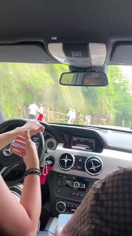 A first-person perspective shot from inside a Mercedes-Benz car. The driver's hands are on the steering wheel, and their right hand is gesturing. The car is moving along a road that is flanked by lush green trees and vegetation. On the side of the road, there are several people visible. The road appears to be a coastal road or a scenic route, as suggested by the presence of water in the distance. The weather is bright and sunny. The dashboard of the car, including the infotainment system, air vents, and steering wheel, is clearly visible. The interior of the car is dark, while the exterior is brightly lit. A rearview mirror shows the reflection of a person's face. The overall scene suggests a leisurely drive through a scenic landscape.