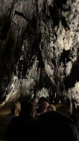 A low-angle interior shot shows the back of several people seated on yellow benches, moving slowly through a dimly lit cave. The camera is positioned behind the seated individuals, looking forward into the cave. Above them, the cave ceiling is high and uneven, displaying dramatic rock formations in shades of dark gray, black, and lighter white or pale gray, illuminated by unseen artificial lights. Some stalactites are visible hanging from the ceiling. The people are mostly silhouetted, with some details of their heads and shoulders visible. The movement suggests they are on a guided tour, possibly on a tram or train.