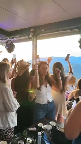 A group of people are dancing and holding up their phones to record at an outdoor event. They are positioned on a raised platform or balcony, with a scenic view of mountains and a body of water in the background. The sky is overcast with a sunset glow. There are speakers and lighting equipment visible, suggesting a DJ or live music performance is taking place. The people are dressed in casual summer attire. Some are holding drinks. The overall atmosphere appears to be festive and lively.