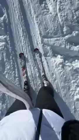 The video shows a person skiing uphill using a ski lift. They are wearing skis, dark pants, a white jacket, a black helmet, and blue goggles. The person is holding onto a gray pole for support as they are pulled uphill on a snow-covered slope. The sun is shining, and the sky is blue. There are tracks in the snow and some trees visible in the distance. The video is filmed from the person's point of view.