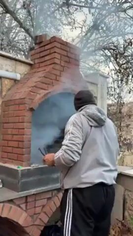 A man wearing a gray jacket, black sweatpants, and a black balaclava tending a fire in a red brick outdoor oven. He holds tongs and is moving pieces of burning material. White smoke billows from the oven. The oven sits on a base constructed from concrete blocks. The surroundings include foliage and part of a fence.