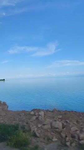 The video showcases a scenic view of a vast, calm body of water under a clear blue sky dotted with wispy white clouds. The water is a serene blue, reflecting the sky and appearing almost glassy. In the foreground, a rough, earthy terrain with scattered rocks and some sparse vegetation suggests a natural, undeveloped shoreline.

On the left side of the frame, a distant shoreline is visible, featuring a patch of sand, a few trees, and what appear to be buildings, hinting at a settlement or resort area. A couple of small figures can be seen on the sandy beach, suggesting the presence of people enjoying the outdoors. The overall atmosphere is tranquil and peaceful, evoking a sense of open space and natural beauty.

The lighting and clear skies indicate it is likely daytime, possibly late morning or early afternoon, with good weather conditions. The visual cues suggest a lakeside or seaside location. The motion in the video implies the viewer is either moving along the shoreline or observing the scene from a slightly elevated or moving vantage point. There is no visible text in the media file. The location context of Bulan-Segettu, Kyrgyzstan, points to a potentially remote and natural landscape.