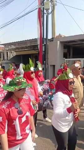 A group of people, mostly women, are walking in a procession on a sunny day. They are wearing red and white outfits, with some individuals sporting decorative headbands with green leaves. Many of the women are wearing red headscarves. One woman in the foreground is wearing a camouflage-patterned hat and a red shirt with white lettering. Another person is carrying a banner with red cloud-like patterns and text that appears to be in Indonesian. The procession is taking place on a street lined with houses and utility poles. The overall atmosphere suggests a celebratory or commemorative event.
