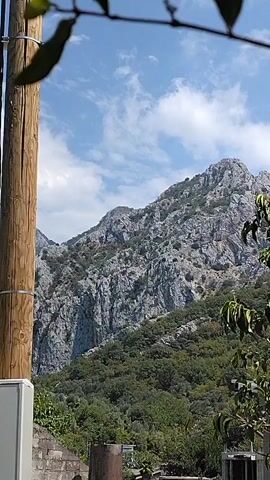 The video shows a view of a rocky mountain with lush green trees in the foreground. The sky is blue with some white clouds. There are some buildings and a wooden pole visible in the lower part of the frame. The camera appears to be stationary.