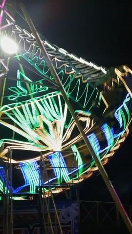 The video shows a Ferris wheel, brightly lit with colorful lights, turning at night. The wheel is decorated with green, yellow, and blue lights. It is partially obscured by a structure of yellow poles and blue and white panels. A man wearing a hat and dark shirt stands on the left, near the base of the Ferris wheel. The background is dark, with trees visible in the distance. The camera angle is looking up at the Ferris wheel.