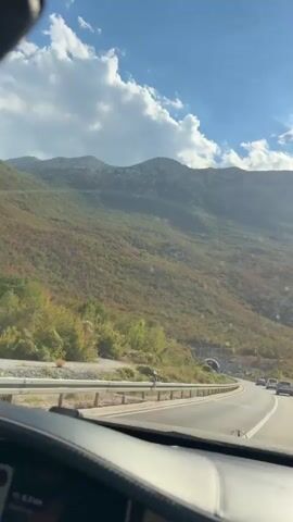 A POV shot from inside a vehicle shows a highway winding through a mountainous landscape under a bright, cloudy sky. The road has multiple lanes separated by white lines and a metal guardrail runs alongside it. In the distance, a tunnel entrance is visible, with a few cars driving towards it. The mountains are covered in trees and vegetation, with some areas appearing to be autumnal. The sun is shining brightly, casting lens flares across the windshield. The dashboard of the vehicle is visible in the foreground.