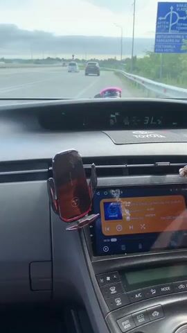The video is filmed from the perspective of a passenger inside a Toyota car, looking at the dashboard and the road ahead. The car is moving on a highway. In the foreground, a phone mount with a red phone is visible on the dashboard. To the right, the car's infotainment screen displays music playback. Further to the right, prayer beads with a crucifix are hanging from the rearview mirror. The road ahead shows other vehicles in motion. The sky is partly cloudy. A speed limit sign is partially visible on the right side of the road.