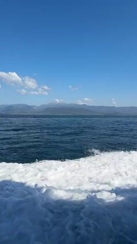 The video shows a view from a boat moving through the water. The water in the foreground is churning white foam, indicating the boat's wake. The water further out is a deep blue, leading to a distant shoreline with mountains under a clear blue sky with scattered white clouds. The camera appears to be positioned at the stern of the boat, looking backwards. There are no people visible in the frame. The overall impression is one of travel and open water.