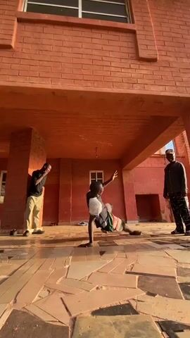 Three young males are present in an outdoor, covered area, likely a veranda or underpass of a building in Kuru, Nigeria. The structure is made of distinctive reddish-brown bricks, and the ground is covered with a tiled, reddish-brown floor featuring an irregular, mosaic-like pattern. The scene is illuminated by warm, directional sunlight, suggesting either late afternoon or early morning.

In the center, one young man, wearing a black and white t-shirt and patterned shorts, is performing breakdancing moves. He transitions through various floor work, including sitting with legs extended, getting up, and executing a move that involves balancing on his hands, showcasing his agility and strength.

Two other young men observe the dancer. On the left, a young man in a dark t-shirt and light-colored pants stands casually, watching the performance. On the right, another young man, dressed in a dark hoodie and patterned pants, stands further back, also attentively observing the dance. A small, white-framed window is visible in the brick wall behind them, and a light fixture hangs from the ceiling of the covered area. The overall atmosphere is focused on the dance performance.
