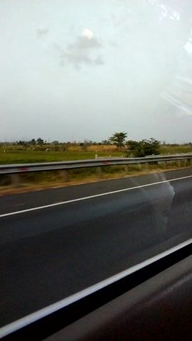 The video shows a view from a vehicle traveling on a highway. The perspective is from the side window, with the road and surrounding landscape visible. The road appears to be a multi-lane highway with a clear separation between lanes indicated by white lines. To the left of the road, there is a grassy area with scattered trees, some of which appear to be palm trees. The sky is overcast, suggesting cloudy weather. The right side of the frame shows a dark, possibly reflective surface, which is likely part of the vehicle's interior or window frame. There are no people or other vehicles clearly visible in the frame. The motion of the vehicle creates a sense of movement along the highway.