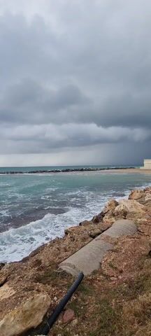 The video shows a seascape under an overcast sky. The sea is a turquoise color, with white foamy waves crashing onto a rocky shoreline.  Further out, a line of rocks acts as a barrier, creating smaller waves closer to the beach.  The sky is filled with thick, grey clouds. On the right side of the frame is a concrete pipe and some rocks. The overall tone is bleak and somewhat stormy. There are no people visible in the scene.